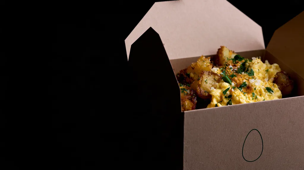 Soft scrambled eggs with crispy golden potatoes, herbs and parmesan inside a folded kraft fiber box on a black studio background.